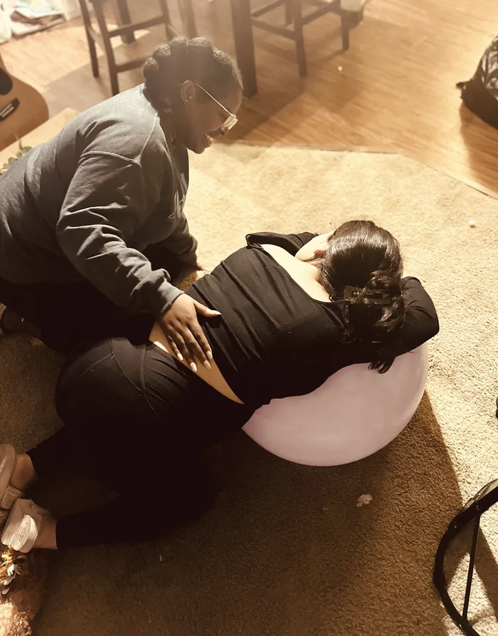 Morgan kneeling beside a laboring mother, applying counter pressure to her lower back as she rests over a birth ball at home.