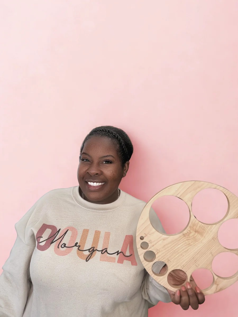 Morgan smiling in a cream Doula sweatshirt, holding a wooden cervical dilation teaching chart.