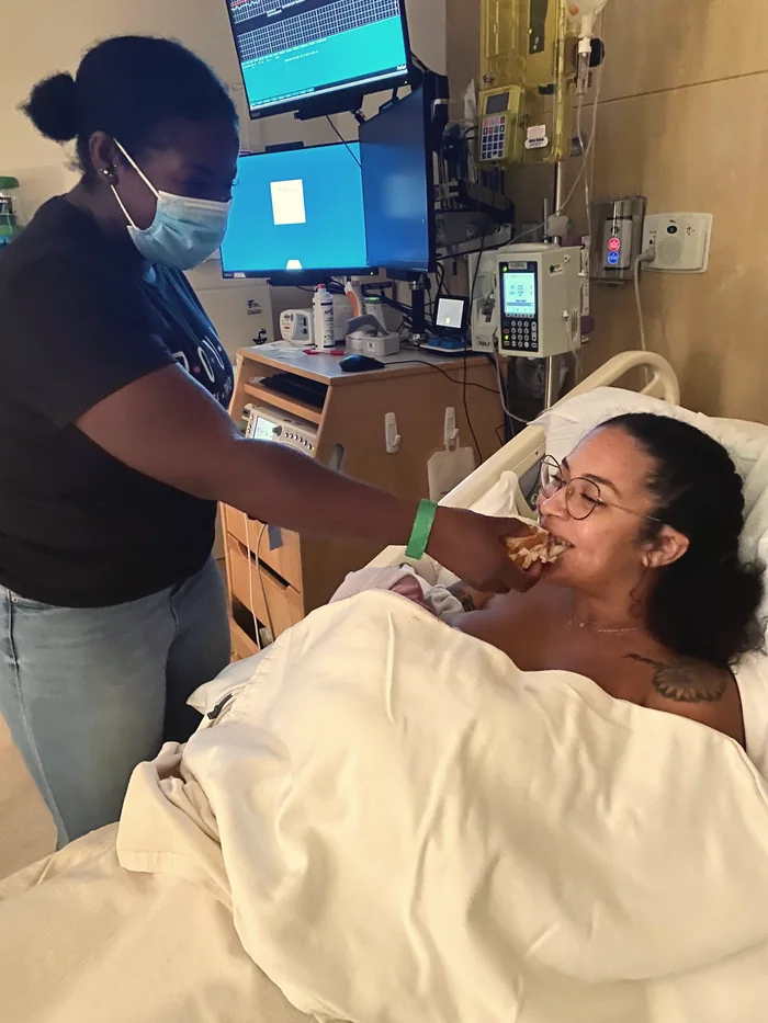Morgan feeding a new mother a sandwich in her hospital bed while she holds her newborn.